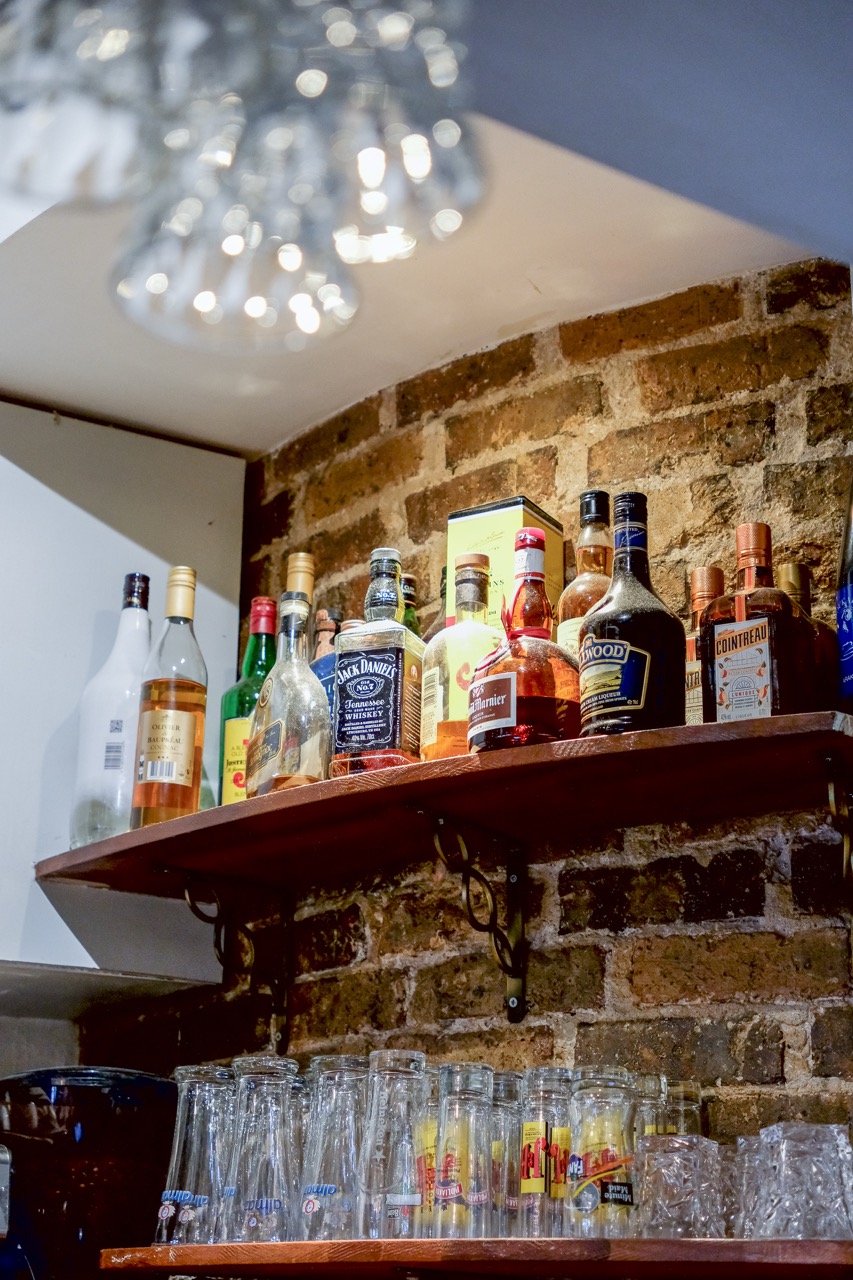 Étagère du bar de Baladna avec bouteilles et verres devant un mur de briques